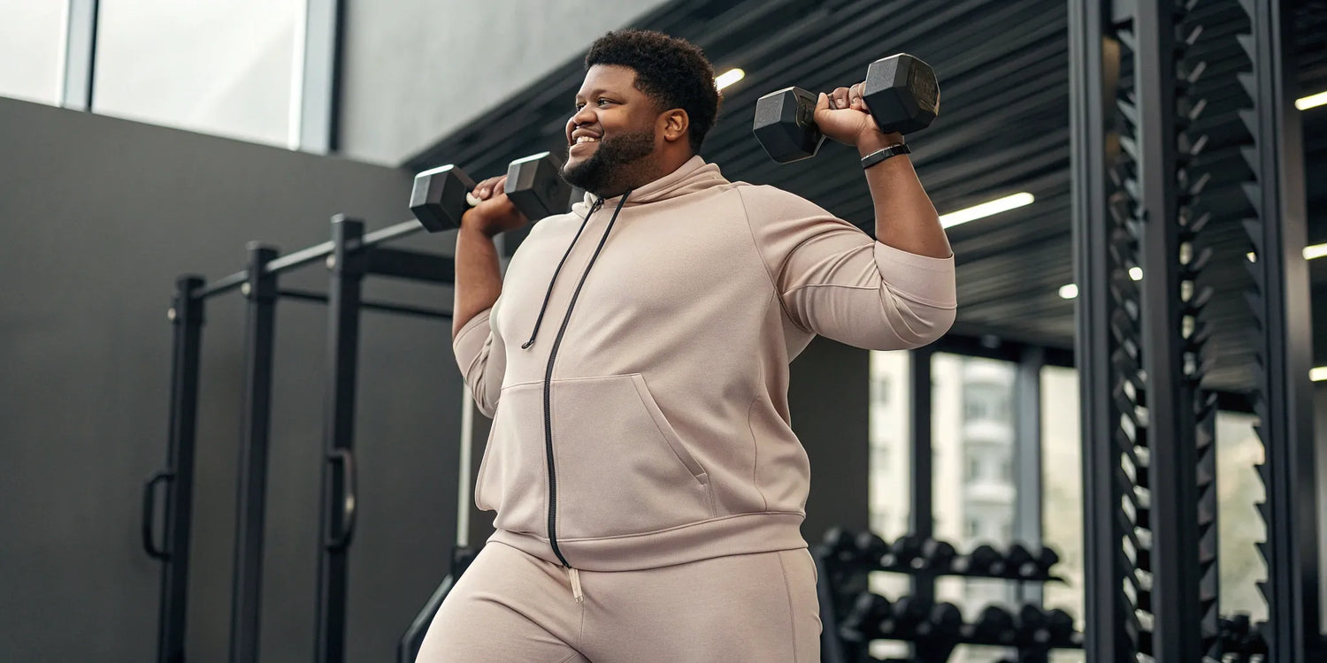 A big guy in well-fitting workout clothes lifts dumbbells at the gym.