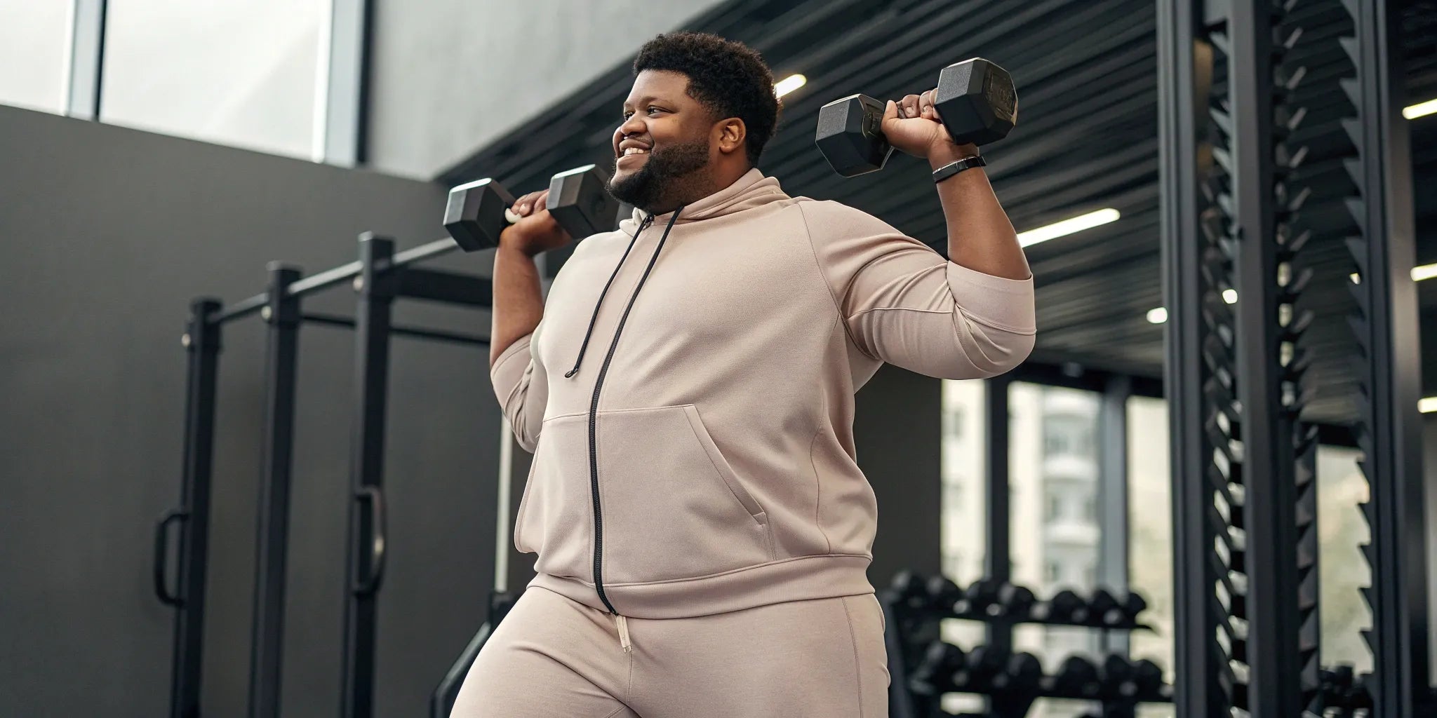 A big guy in well-fitting workout clothes lifts dumbbells at the gym.